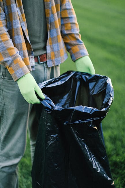 Three large black plastic bags filled with waste materials are placed on a paved sidewalk against a dark metal fence, with dense foliage and trees in the background. The bags are crumpled and appear t