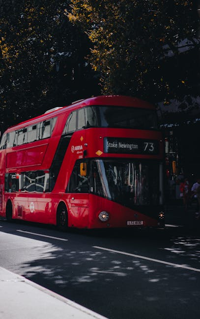 A double-decker bus in bright red livery, parked on a street with the destination display reading 'Stoke Newington' and route number 73. The bus is positioned under a canopy of leafy trees, casting sh